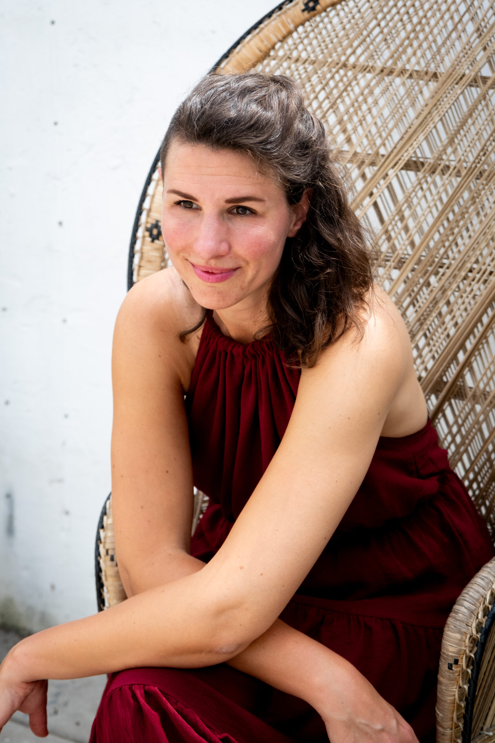 Renée Overboom * Afbeelding van een vrouw in een bordeaurode jurk zittend op een rieten stoel met de armen gekruist over haar benen.